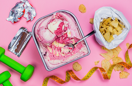 Overhead image of ice cream, chips, chocolate wrappers, hand weights and a measuring tape on a pink table, illustrating the concept that eating disorders can affect your identity, self-worth and self-esteem