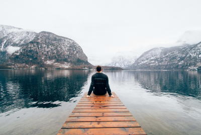 Back view of a man sitting on a long pier jutting out into a large body of water surrounded by mountains, illustrating that you do not have to continue to suffer from depression, there is help.