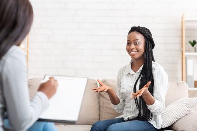 Smiling young black woman sitting on a couch speaking with a therapist, illustrating that ​psychotherapy can be helpful for a wide range of mental health issues, including anxiety, depression, trauma, eating disorders, substance abuse, and relationship problems.