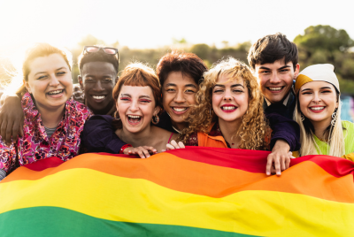Racially and gender diverse group of smiling young people standing behind a pride rainbow flag, illustrating the concept that at Healing Path Therapy, we have therapists who understand the complexities of being 2SLGBTQIA+