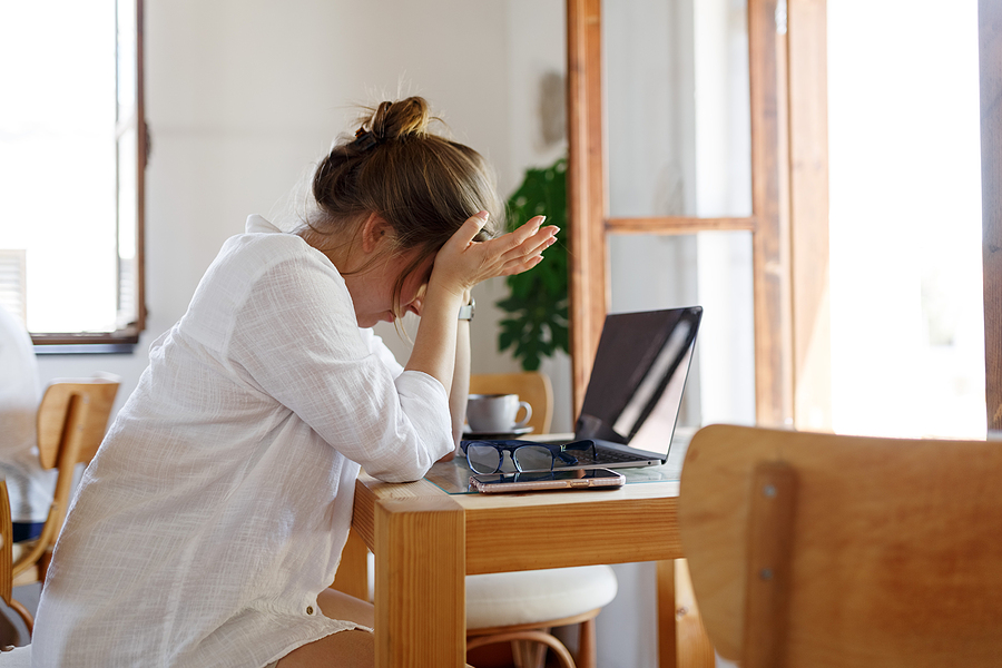 Image of young caucasian woman at desk in front of laptop with head leaning against her hands, illustrating the concept that stress from work can compound the "regular" stress we experience in life.