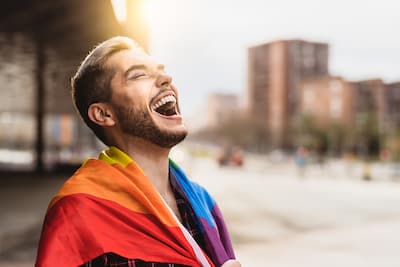 Laughing Caucasian man wrapped in a rainbow flag, illustrating the concept that gender identity is complex, personal, and deeply rooted in how individuals perceive themselves.