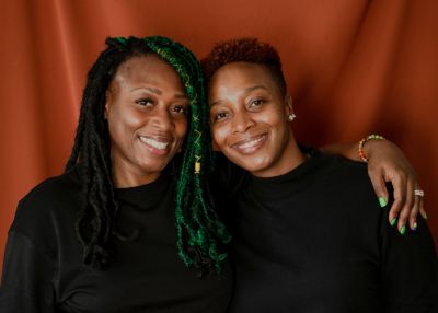 Image of two smiling women of color with arms around each other, illustrating that therapy can help couples navigate the complexities of their relationship.
