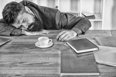 Black and white image of a bearded young man asleep on his arm at a table with books illustrating the concept that Burnout manifests as complete emotional, physical and mental exhaustion.
