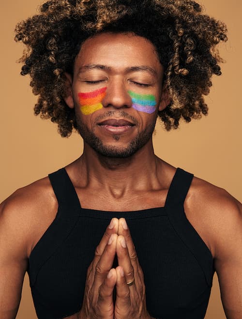 Non-binary person of color with rainbow stripes painted under his closed eyes with hands in a "namaste" pose, illustrating the concept that anxiety comes in many sizes and shapes