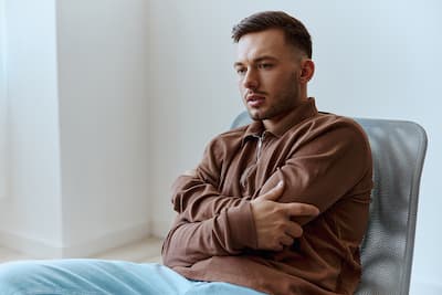 Caucasian young man sitting in chair with arms around himself looking thoughtful, illustrating the concept that therapy can be a transformative experience.