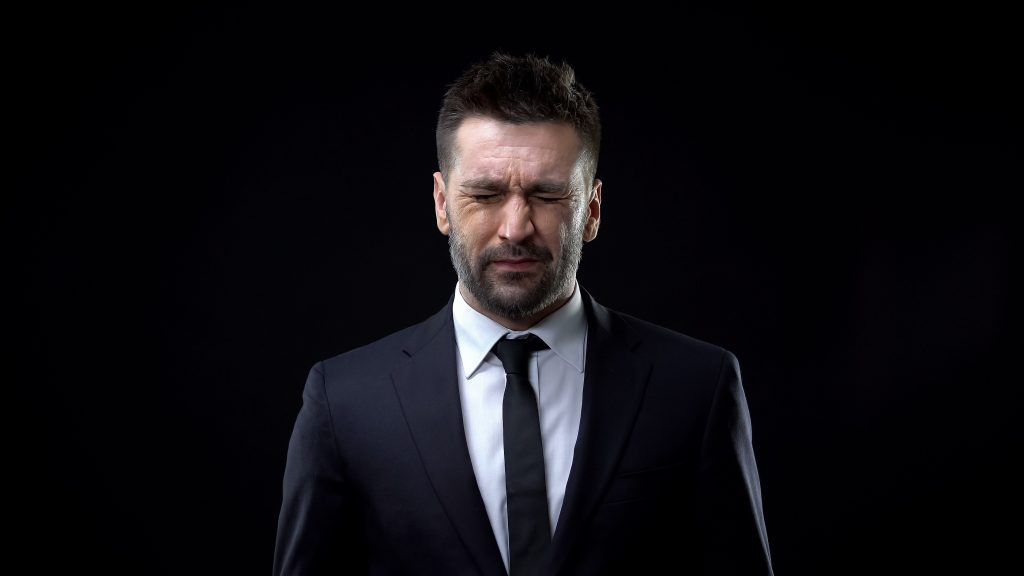 Image of a young Caucasian bearded man in a business suit tightly squeezing his eyes shut against a black background, illustrating that although individuals with narcissistic personality disorder rarely seek treatment, psychotherapy can help these individuals learn how to better relate to others and strengthen relationships, as well as understand their emotions and control their impulses.