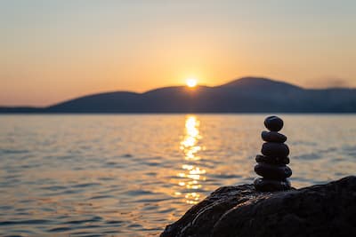 Stack of pebbles on a large rock in front of a large body of water at sunset, illustrating the concept that our anxiety therapy group can help you manage and overcome anxiety.