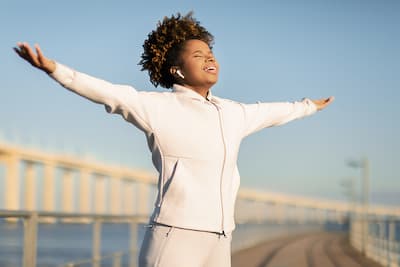 Image of happy young black woman in a jogging suit with eyes closed and arms out wide illustrating that therapy can help you become the best version of yourself.