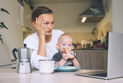 Caucasian young woman sitting at kitchen table in front of laptop with baby on her lap illustrating the concept that there are many reasons which cause stress.